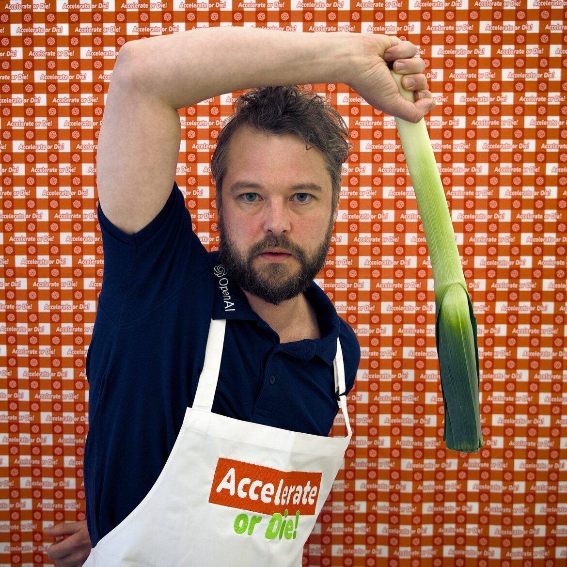 Florian Egermann wearing a dark blue shirt and a white apron with the text "Accelerate or Die" stands against a backdrop with repeated logos. He holds a leek over his head with his left hand, looking intently at the camera.