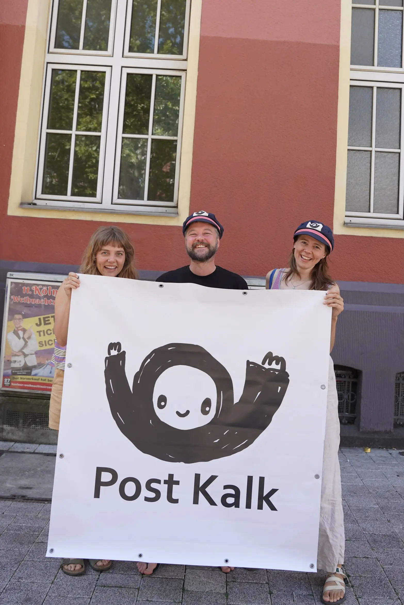 Three smiling people stand outside a red building holding a large white banner showing a hand-drawn dark figure with a round smiling face and raised arms, above the words 'Post Kalk'.